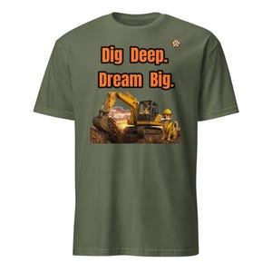 Op de afbeelding: Een olijfgroen t-shirt met een afbeelding van een gele graafmachine en een hond met een gele veiligheidshelm en een reflecterend vest. De tekst "Dig Deep. Dream Big." staat in het oranje erboven, met een klein pootafdruk-logo.