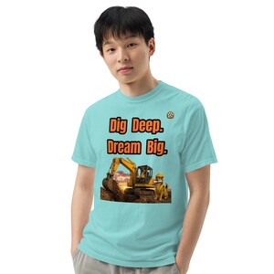 Op de afbeelding: Een lichtblauw t-shirt met de tekst Dig Deep. Dream Big. in oranje letters boven een afbeelding van een gele graafmachine die in de aarde graaft, met een hond met een gele veiligheidshelm in een landschap bij zonsondergang.