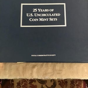 Könnte beinhalten: Eine dunkelblaue, rechteckige Aufbewahrungsbox mit weißem Text, der 25 Years of U.S. Uncirculated Coin Mint Sets und Postal Commemorative Society lautet. Die Box hat einen weißen Rahmen um den Haupttitel und eine glatte, einfarbige Oberfläche.