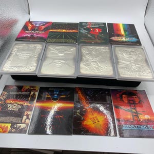 Op de afbeelding: Een verzameling Star Trek filmmemorabilia bestaande uit vier kleurrijke filmposters, vier zilverkleurige reliëf metalen kaarten in beschermhoezen en vier extra filmposters met de titels Star Trek II, III, IV en V.