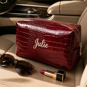May include: A deep red, rectangular cosmetic bag with a crocodile-embossed texture and a gold-tone zipper. The name Julie is embroidered in white script on the front. Beside the bag are dark-rimmed sunglasses and a tube of red lipstick.