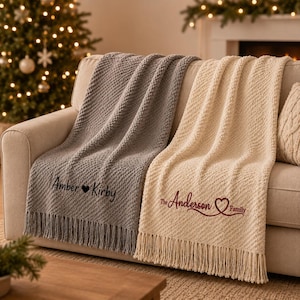 May include: Two textured throw blankets with fringe edges draped over a sofa. The grey blanket features the embroidered text Amber heart Kirby in black. The cream blanket features the embroidered text The Anderson heart Family in dark red.