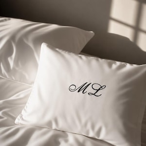 May include: A white rectangular pillowcase featuring black embroidered cursive initials ML centred on the front. The fabric has a smooth finish and is positioned on a bed with another white pillow visible in the background under soft lighting.