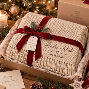 May include: A cream-coloured knit throw blanket with fringe, personalised with the text Amelia + Noah and 08.18.2026. It is wrapped in a deep red velvet ribbon with a tag reading A Warmer Tomorrow and a small sprig of pine and red berries.