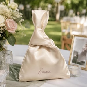 May include: A champagne-colored satin knot bag featuring a triangular silhouette with a single handle loop. The fabric has a smooth, lustrous finish. The name Emma is embroidered in a delicate, cursive script on the lower front of the bag.