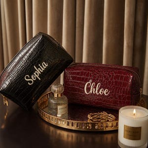 May include: Two rectangular cosmetic bags with a crocodile-embossed texture. One bag is black with the name Sophia embroidered in white, and the other is deep red with the name Chloe embroidered in white. Both feature gold-tone metal zippers.