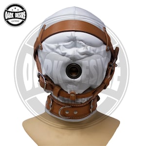 May include: A white padded hood mask featuring a central circular breathing port and a tan leather harness system with silver-toned metal buckles and rivets. The mask includes a matching tan leather collar with a D-ring at the front center.