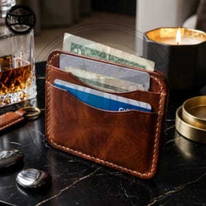 May include: A brown leather card holder featuring visible light-colored stitching along the edges. The wallet has multiple slots containing a paper banknote and two plastic cards, one of which is blue and white, displayed in a vertical orientation.
