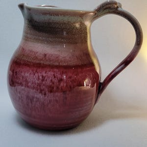 Puede incluir: Una jarra de cerámica con cuerpo redondeado y asa curva. El esmalte transita desde un burdeos profundo y saturado en la base hasta un tono malva y gris moteado más claro cerca del borde, mostrando una sutil textura agrietada.