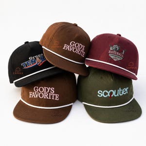 May include: A collection of five baseball caps in black, brown, maroon, and olive green, each featuring a white rope detail across the brim. The caps display various embroidered text and logos, including 'GOD'S FAVORITE', 'Grupo TENARI', 'BASEBALL CRIMSON PRIDE', and 'scouter'.