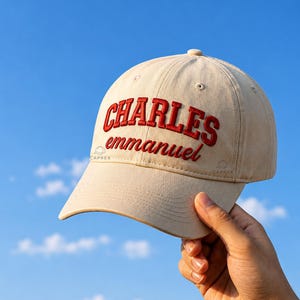 May include: A beige baseball cap featuring the text CHARLES emmanuel embroidered in red thread on the front panel. The cap has a curved brim, a structured crown with ventilation eyelets, and a button at the top centre against a blue sky.