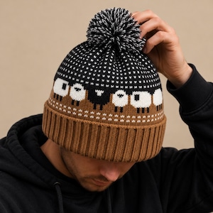 May include: A knit beanie featuring a brown ribbed cuff and a black upper section with a white sheep pattern. The hat is topped with a black and white pom-pom. The design includes a row of white and black sheep figures against a dark background.