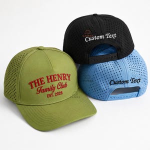 May include: Three baseball caps with perforated side and back panels. One olive green cap features red embroidered text reading THE HENRY Family Club EST. 2026. One black cap and one light blue cap both feature the embroidered text Custom Text.