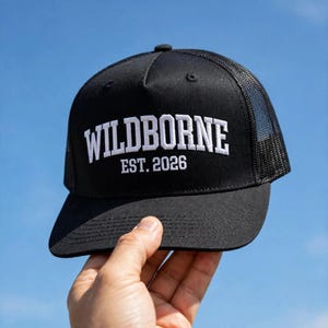 May include: A black trucker cap featuring a mesh back and a solid front panel. The front displays white embroidered text that reads WILDBORNE in a bold, arched font, with the text EST. 2026 positioned directly underneath in a smaller font.