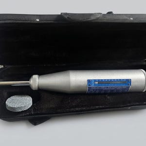 May include: A silver concrete test hammer with a blue calibration chart and a sliding indicator, positioned inside a black zippered carrying case. A small, circular gray abrasive stone is included in the case next to the testing instrument.