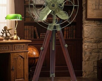 May include: A floor-standing electric fan featuring four pale green blades encased in a circular metal wire cage. The fan is mounted on a dark brown wooden tripod stand with brass-colored hardware and a central chain connecting the three legs.