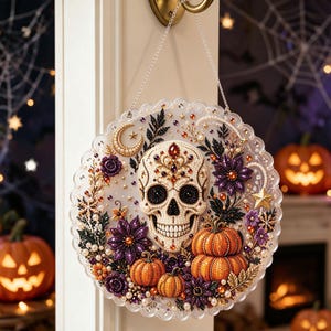 May include: A circular wall hanging featuring a central skull decorated with beads and gems, surrounded by orange pumpkins, purple flowers, and a crescent moon. The design is embellished with small pearls and stars on a scalloped, translucent base.