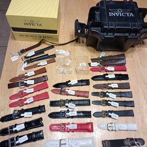 May include: A collection of watch straps in various colors and materials, including leather and fabric, with metal buckles. The straps are laid out on a wooden surface, alongside an Invicta watch box and a black Invicta watch case.