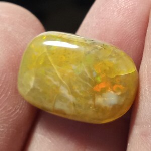 May include: A yellow opal gemstone with a smooth, rounded shape. The opal has a white and orange iridescent sheen.