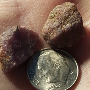 May include: Two rough, reddish-purple gemstones next to a U.S. dime coin. The gemstones have a textured surface and varying shades of purple and brown. The coin shows the profile of a man and the year 2017.