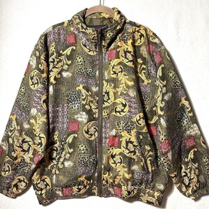 Könnte beinhalten: Eine langärmelige Reißverschlussjacke mit olivgrüner Basis und einem sich wiederholenden Muster aus goldenen Akanthusblättern, schwarz-weißen Tierdruckquadraten und kleinen roten kreisförmigen Motiven. Die Jacke hat einen Stehkragen und einen durchgehenden Reißverschluss.