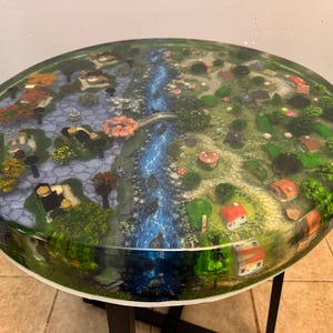 Può includere: Un tavolo rotondo con un piano in resina lucida che presenta un dettagliato paesaggio in miniatura. La superficie mostra un fiume blu centrale che scorre tra due lati distinti, uno con sentieri in pietra e alberi, l'altro con piccole case.