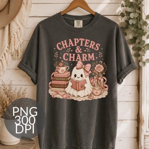 May include: A charcoal gray t-shirt featuring a graphic of a white ghost wearing a pink plaid bow and reading a book. The text reads "CHAPTERS & CHARM" above the ghost, surrounded by stacks of books, floral arrangements, and tea cups in pink tones.