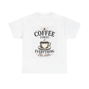 May include: A white short-sleeve t-shirt featuring a central graphic of a coffee cup on a saucer. Above the cup, the text reads COFFEE FIRST. Below the cup, the text reads EVERYTHING Else Later. A small coffee bean icon is positioned above the text.
