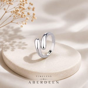 May include: A polished silver-toned bypass ring featuring two smooth, rounded teardrop-shaped ends that do not meet. The ring has a reflective metallic finish and is displayed on a circular stone-like base with the text TIMELESS ABERDEEN.