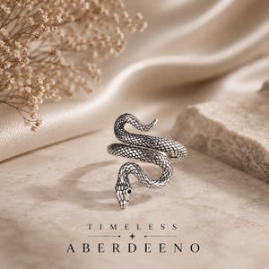 Può includere: Un anello in metallo color argento a forma di serpente avvolto con texture a scaglie dettagliate e piccoli occhi neri. Il corpo del serpente si avvolge per formare la fascia dell'anello. Il testo TIMELESS ABERDEENO è stampato in basso.
