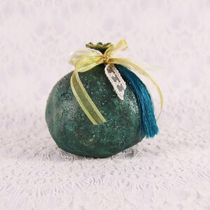 Peut inclure: Un objet décoratif rond et texturé en forme de grenade, de couleur vert foncé. Il est orné d'un ruban doré, d'un morceau de dentelle blanche et d'un pompon bleu sarcelle profond. La surface présente une finition métallique et mouchetée.