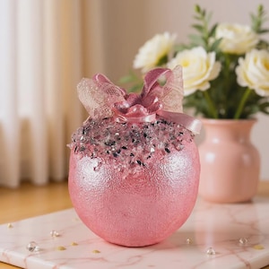 Peut inclure: Une décoration sphérique rose avec une finition métallique texturée, ornée d'une bande de fragments de verre concassé transparents et argentés sur le dessus. L'objet est surmonté d'un nœud décoratif en dentelle rose pâle et ruban satiné.