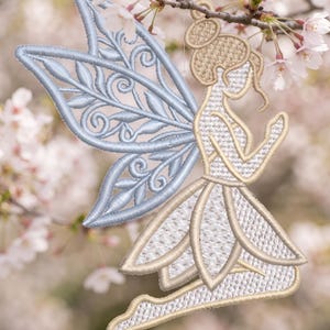 May include: An embroidered ornament shaped like a kneeling figure with large, light blue wings featuring intricate leaf and scroll patterns. The figure has a gold-toned body with a textured stitch pattern, a bun hairstyle, and a skirt with petal-like layers.