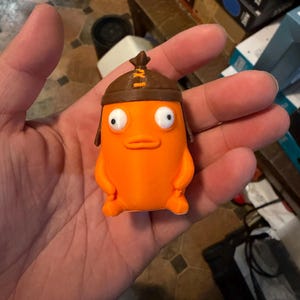 May include: A small, bright orange figurine with a smooth, rounded body, two white eyes with black pupils, and a simple mouth. The character wears a brown, rounded cap with a top knot and orange stitching details on the front of the headpiece.