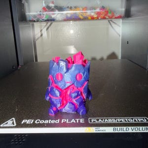 Peut inclure: Une figurine imprimée en 3D présentant un extérieur violet foncé semblable à de la roche, avec des structures cristallines rose vif dépassant du sommet et visibles à travers des fissures profondes dans le corps. Elle a deux yeux roses circulaires.