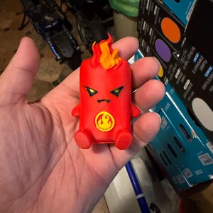 Peut inclure: Une petite figurine rouge imprimée en 3D représentant un personnage stylisé avec une touffe en forme de flamme sur la tête. Elle présente des yeux jaunes anguleux, une petite bouche et un emblème jaune circulaire sur la poitrine.