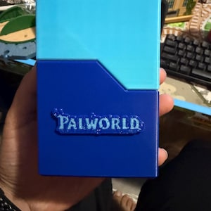 Könnte beinhalten: Eine rechteckige Deck-Box mit zweifarbigem Design, bestehend aus einem hellblauen oberen Teil und einem dunkelblauen unteren Teil. Auf der Vorderseite befindet sich der Schriftzug Palworld in erhabenen, texturierten Buchstaben.