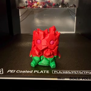 Może przedstawiać: Figurka wydrukowana w technologii 3D o fasetowanej, przypominającej skałę teksturze. Górna część jest jaskrawoczerwona z dwoma okrągłymi różowymi oczami, a dolna część jest jaskrawozielona. Obiekt stoi na czarnej płycie pokrytej powłoką PEI.