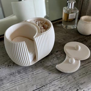 Op de afbeelding: Een witte, geribbelde organizer met twee compartimenten voor badkamerbenodigdheden. De ene kant bevat wattenstaafjes, de andere kant wattenschijfjes. De houder heeft een bijpassend, tweedelig afneembaar deksel met kleine ronde knopjes.