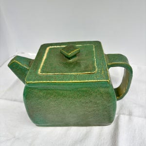 May include: A square-shaped ceramic teapot with a textured, mottled green glaze and a matching lid. The teapot features a prominent spout on the left side, a sturdy handle on the right, and a small, square knob centered on the lid.