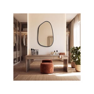May include: An asymmetrical wall mirror with a thin black frame mounted above a wooden vanity table. The vanity features three drawers, a set of makeup brushes, perfume bottles, and a small box. A round, rust-coloured velvet ottoman sits below.