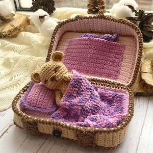 May include: A crocheted suitcase in beige and brown containing a small tan bear toy tucked under a purple and pink variegated blanket. The interior features a pink crocheted pillow and a purple pocket, with a dark button closure on the exterior.