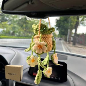 May include: A crocheted hanging plant ornament featuring a tan pot, green leaves, and white flowers with yellow centers, suspended by a tan cord. A small brown cardboard box with the text 96CRVFT printed on the side is visible in the corner.