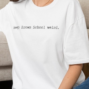 May include: A white short-sleeved t-shirt featuring the black printed text keep Brown School weird. centered on the chest. The garment has a crew neckline and a relaxed fit, showcasing a simple design with clear, typewriter-style lettering.