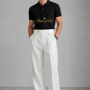 Könnte beinhalten: Ein schwarzes kurzärmeliges Poloshirt mit Kragen, drei Knöpfen und dem gelben Schriftzug Dapper EraCo mit Kronensymbol. Dazu wird eine weiße Hose mit hoher Taille, weitem Bein, geknöpftem Bund und schwarzen Schuhen kombiniert.