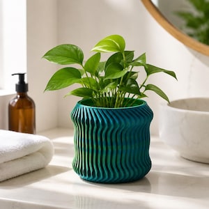 Puede incluir: Una maceta de color verde azulado vibrante con una textura vertical ondulada y acanalada. El recipiente cilíndrico contiene una exuberante planta pothos verde con hojas en forma de corazón. La maceta presenta un borde superior e inferior.