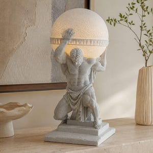 May include: A gray stone-textured table lamp featuring a muscular figure kneeling on a square base while holding a large, spherical, illuminated white globe above their head. The figure wears a draped cloth around the waist and has detailed musculature.