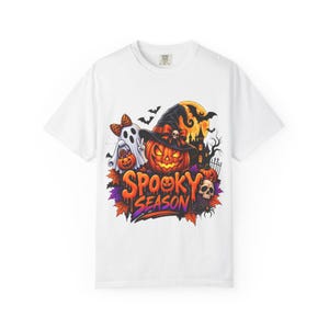 Puede incluir: Una camiseta blanca con un gráfico colorido de Halloween que muestra un fantasma, una calabaza con sombrero de bruja, una calavera, murciélagos y una casa encantada. El texto "SPOOKY SEASON" aparece en letras naranjas y moradas debajo.