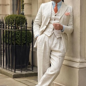 May include: A three-piece cream linen suit consisting of a blazer, a button-up vest, and pleated trousers. The suit is worn with a light blue dress shirt, an orange necktie, a light pink pocket square, and two-tone white and brown leather shoes.