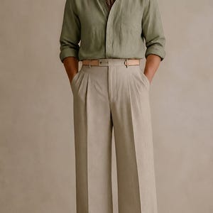 May include: A sage green long-sleeved button-down shirt with a collar, a patterned neck scarf, beige pleated trousers with a wide-leg cut, a light tan leather belt, and brown leather shoes. The outfit features a neutral color palette throughout.